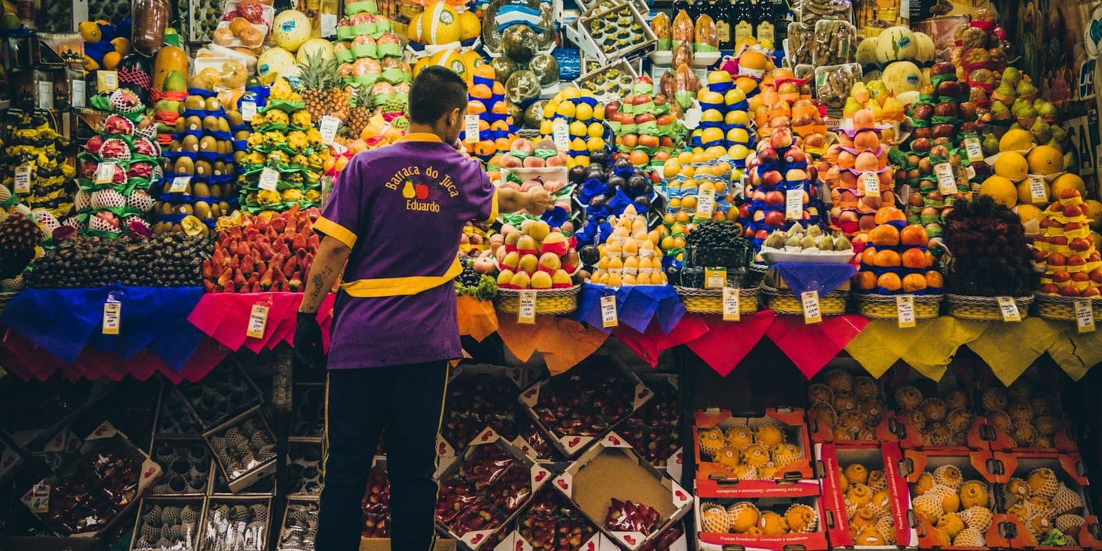 Feirante de costas diante de uma banca de frutas, com as pilhas de frutas subindo do balcão até o teto.