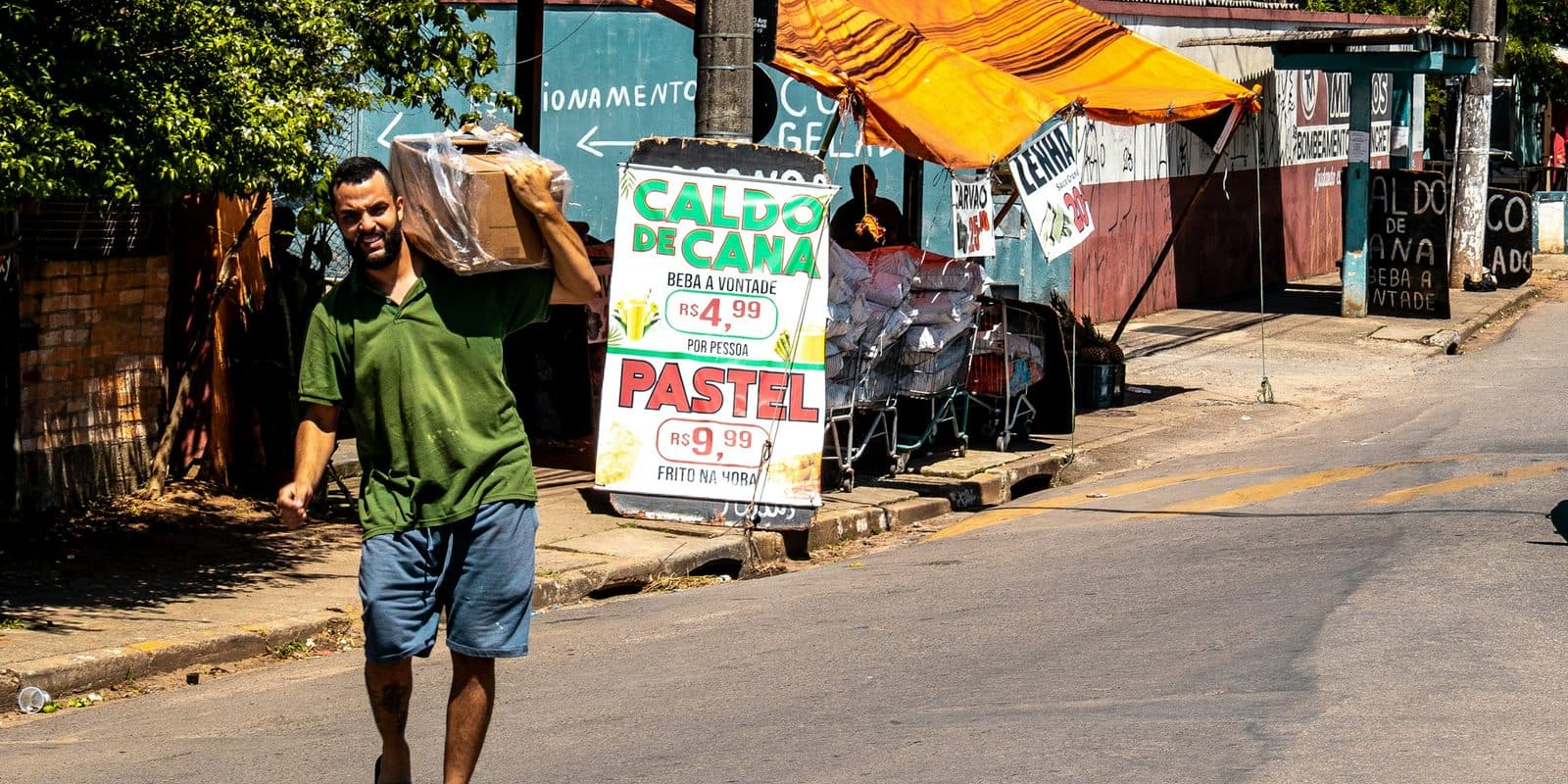 Homem atravessa uma rua carregando uma caixa no ombro, ao lado de uma barraca de caldo de cana e pastel.
